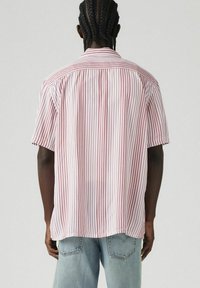 Camicia a maniche corte con strisce verticali rosse e bianche, colletto a bottoni, vestibilità ampia e texture del tessuto liscia. Jeans visibili sotto la camicia.