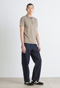 Beige polo tričko s černými akcenty, spárované s volnými tmavě modrými džíny a černými teniskami s bílými tkaničkami, na neutrálním pozadí.
