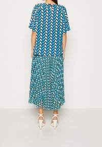 Blå mønstret top med korte ærmer og en nøglehulslukning bagpå, parret med en plisseret blå-grøn mønstret nederdel. Båret med hvide højhælede sko.