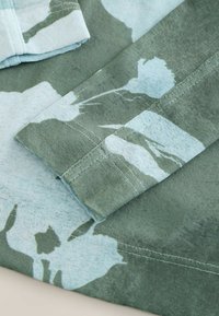 Grön tyg med ljusblå blommönster, visar vikta ärmar och detaljerad sömnad på ett mjukt textil.