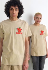 Béžové bavlněné trička s karikaturou jablka s chobotem slona a nápisem "OBEY" v červené barvě, které mají na sobě dvě osoby.