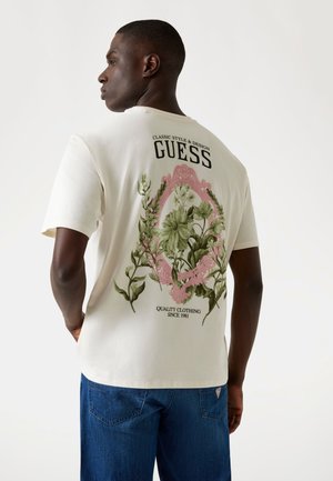 Balta medvilnės marškinėliai su gėlėtos grafikos raštu žalia ir rožine spalvomis ant nugaros, su užrašu "GUESS" viršuje ir "QUALITY CLOTHING SINCE 1981" apačioje.