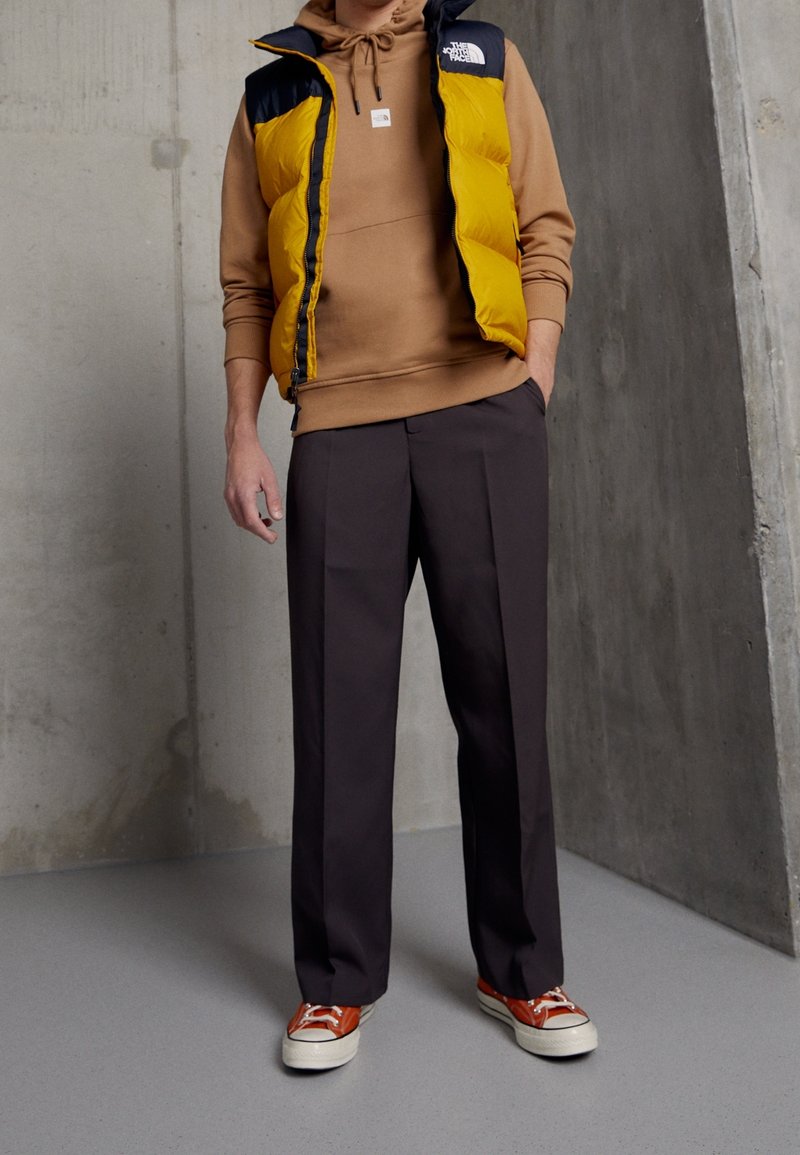 Personne portant un sweat à capuche beige, un gilet matelassé jaune et bleu marine, un pantalon noir et des baskets montantes orange, debout contre un mur en béton gris.