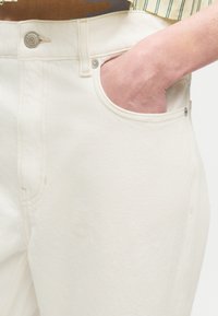 Χέρι στην τσέπη ψηλόμεσου, σχεδόν άσπρου τζιν με μεταλλικό κουμπί και ριγωτό πουκάμισο περασμένο μέσα.