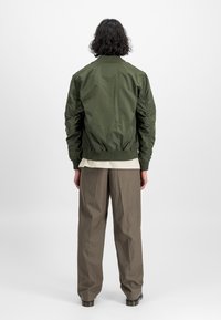 Casaco bomber verde-oliva com gola canelada, bolsos com fecho zip e mangas franzidas. Usado sobre uma camisa bege e calças cinzas.