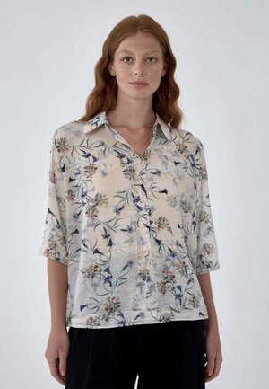 Donna dai capelli rossi che indossa una camicia ampia, abbottonata, con motivo floreale, maniche a tre quarti e pantaloni neri su sfondo neutro.