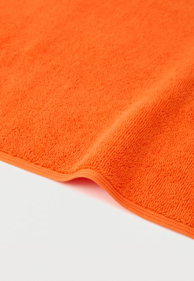 Φωτεινή πορτοκαλί πετσέτα από πετσετέ ύφασμα με ορατή απαλή υφή και ελαφρώς καμπύλη άκρη σε απλό λευκό φόντο.