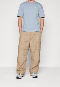 Světlá modrá polokošile s krátkým rukávem a malým logem, spárovaná s volnými béžovými cargo kalhotami a šedými teniskami s oranžovými akcenty.