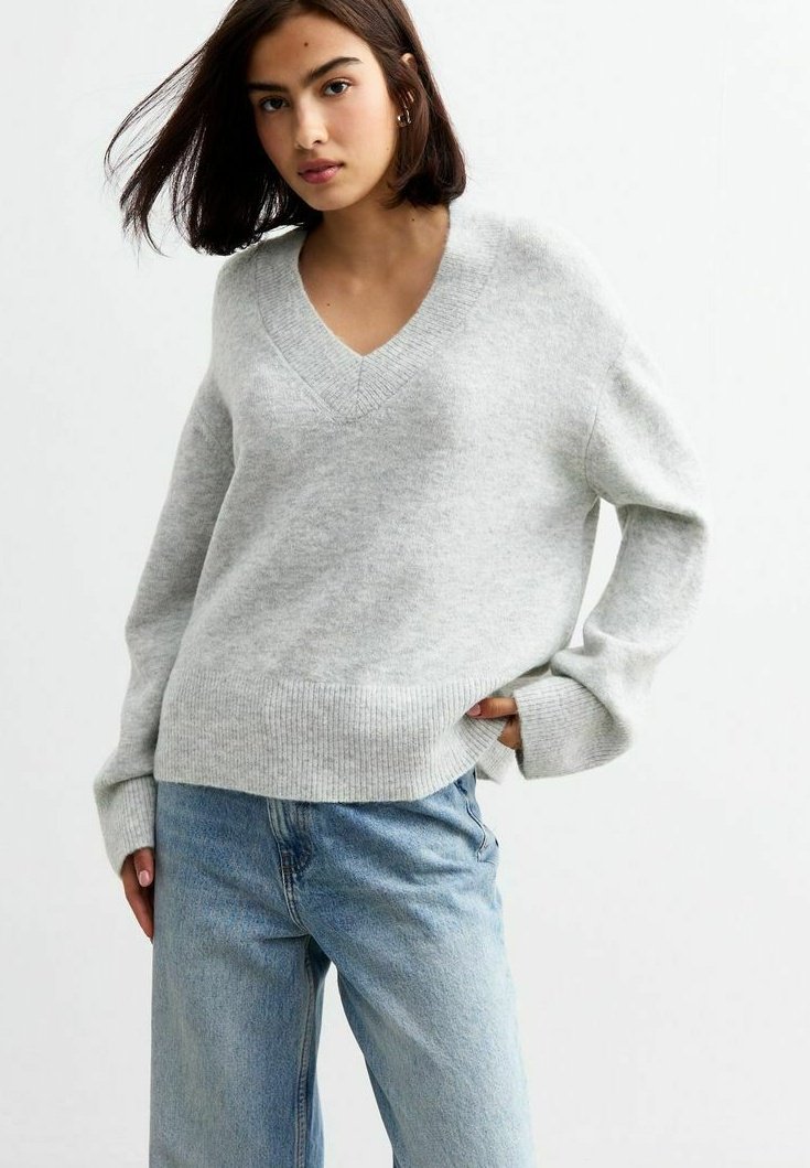 Pull en mélange de laine gris clair, avec un col en V, des manches longues et un ourlet côtelé. Associé à un jean bleu clair, offrant une coupe décontractée.