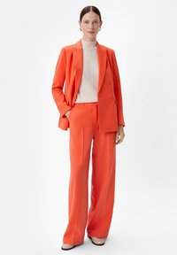 Pomarańczowa dopasowana marynarka podwójnie zapięta z szerokimi spodniami w tym samym kolorze. Jasnobeżowy golf. Proste akcenty biżuteryjne. Gładka faktura materiału.