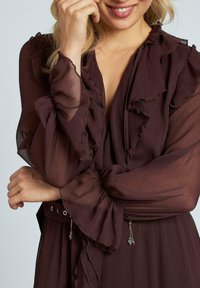 Burgunderblus med genomskinligt tyg, volangkrage och ärmar, midja med bälte som har öglor och fransdetaljer, mjuk textur.