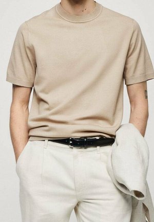 Uomo che indossa un maglione beige a maniche corte infilato in pantaloni crema con cintura nera, tiene una giacca beige chiaro nella mano sinistra.