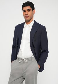 Μπλε μπλέιζερ διπλής σειράς με μαύρα κουμπιά, συνδυασμένο με λευκό πουκάμισο και γκρι παντελόνι με κορδόνι στη μέση. Μεταξένιο ύφασμα, εφαρμοστή ραφή.