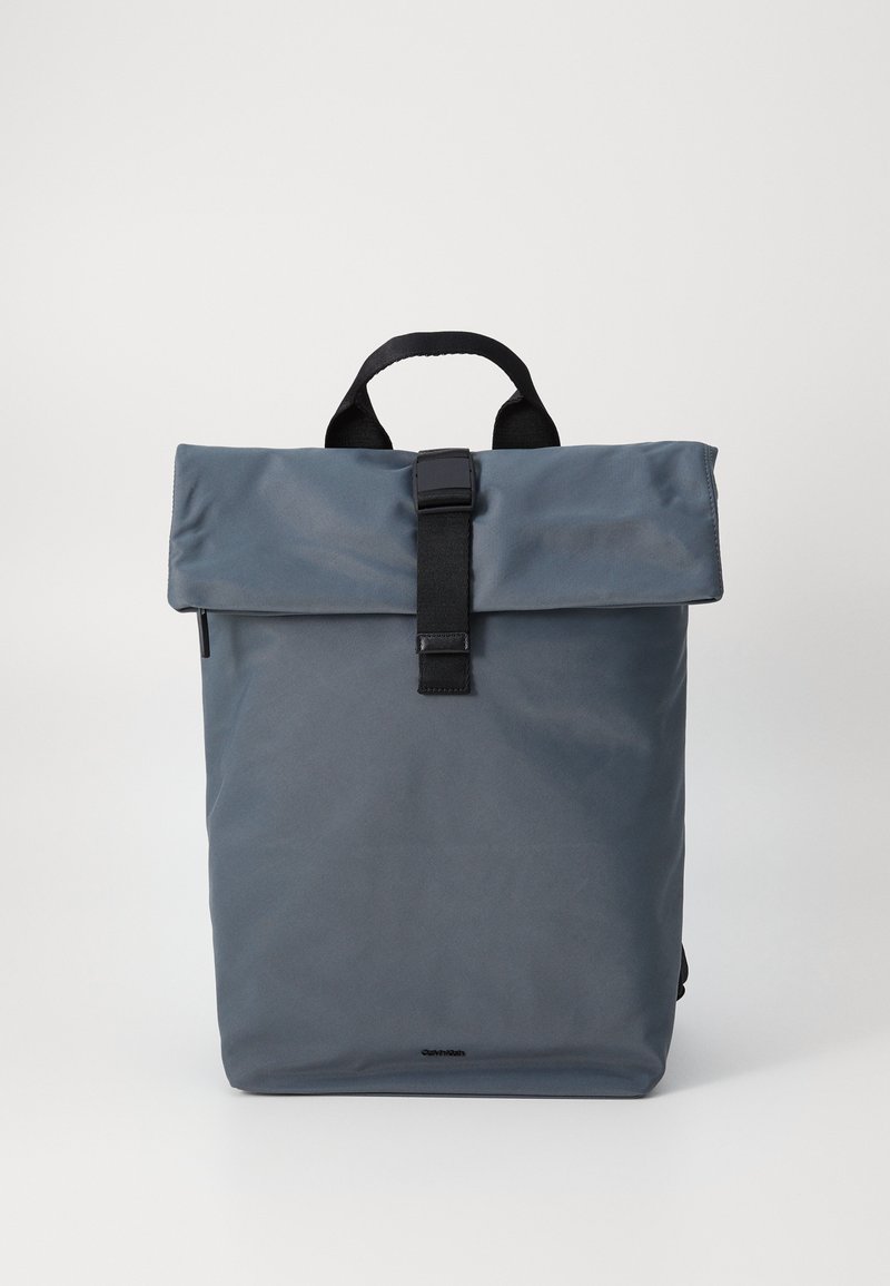 Sac à dos gris à rabat fabriqué en tissu résistant, avec une fermeture à sangle noire et une poignée sur le dessus. Design épuré et minimaliste sans logos visibles.