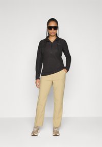 Donna con capelli intrecciati indossa un maglione nero con zip, pantaloni beige, scarpe da ginnastica beige e occhiali da sole neri grandi, in piedi contro uno sfondo semplice.