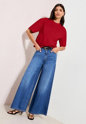 Kobieta w czerwonym krótkim topie, szerokich niebieskich dżinsach z czarnym paskiem i czarnych sandałach na wysokim obcasie, pozująca na tle jasnej ściany.