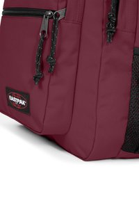 Bordeaux ryggsäck med svarta dragkedjor och snören, strukturerade sidofickor i nät och en svart tyglogo med röda detaljer.