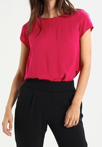 Blusa fucsia de manga corta con cuello redondeado, combinada con pantalones negros de cintura alta que presentan detalles de pliegues en la parte frontal.
