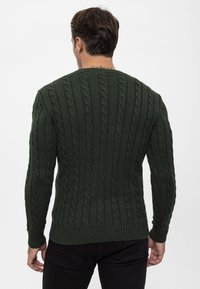 Man i en mörkgrön kabelstickad tröja och svarta byxor, stående med ryggen mot kameran mot en neutral bakgrund.
