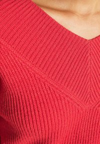 Czerwony prążkowany sweter z dekoltem w serek, pionową fakturą i ukośnym wzorem ściegu na klatce piersiowej. Miękka dzianinowa tkanina.