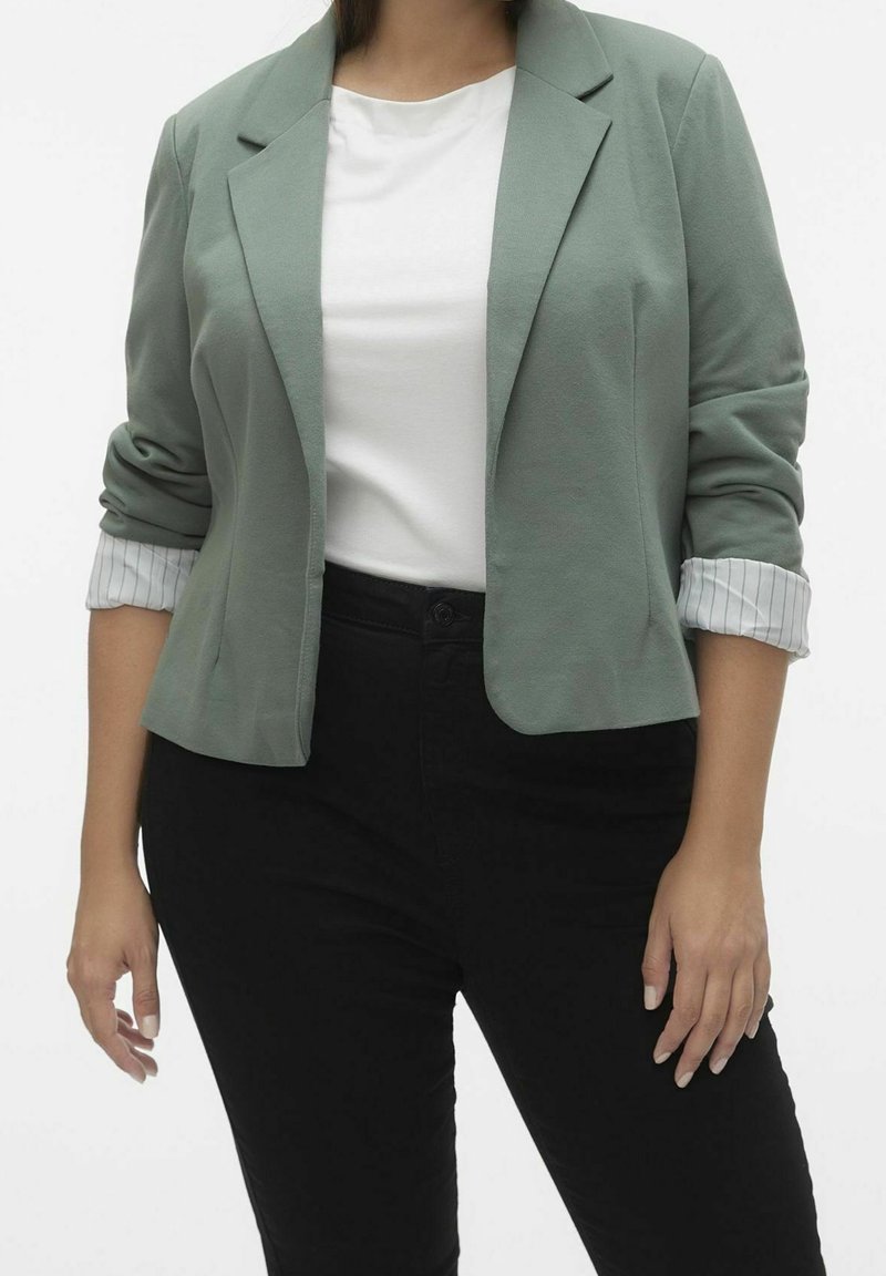 Femme portant un blazer vert clair avec des poignets rayés roulés, un haut blanc et un pantalon noir, sur un fond blanc uni.