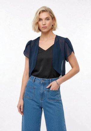 Ung kvinde iført en gennemsigtig marineblå shrug over en sort camisole, højtaljede blå jeans og guld øreringe i hoop-stil, poserer med hånden i lommen.