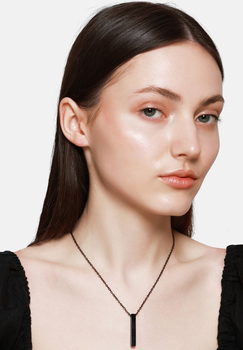 Zwarte halsketting met een rechthoekige hanger, aan een dunne metalen ketting, gedragen met een zwarte top. Het model heeft lang, steil haar en neutrale make-up.