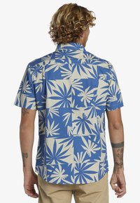 Camisa de manga corta en azul con patrones de plantas blanca. Presenta un cuello clásico y un corte relajado, confeccionada en una tela ligera.