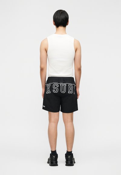 Žmogus, stovintis nugara, dėvintis baltą be rankovių marškinėlį ir juodus šortus su ryškiu balto „KSUBI“ užrašu nugaroje, juodus kojines ir juodus sportbačius.