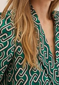 Grüner gemusterter Stoff mit ineinandergreifenden weißen und schwarzen Formen, mit einer glatten Textur. V-Ausschnitt-Design, mit sichtbarem langen Haar daneben.