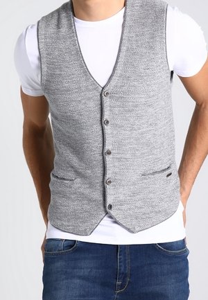 Mies, joka on pukeutunut harmaaseen napitettuun neuleliiviin valkoisen t-paidan ja sinisten farkkujen päällä, seisoo käsineet taskuissa.