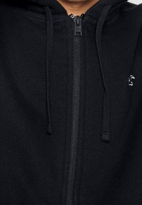 Svart zip-up huvtröja gjord av mjukt material, med justerbara dragsnören, en framtill dragkedja och en liten vit logotyp på bröstet.