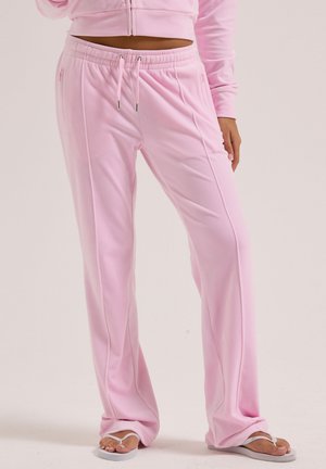 Person trägt hellrosa Sweatpants mit Kordelzug und passende kurze Jacke, steht barfuß in weißen Flip-Flops vor einfarbigem Hintergrund.