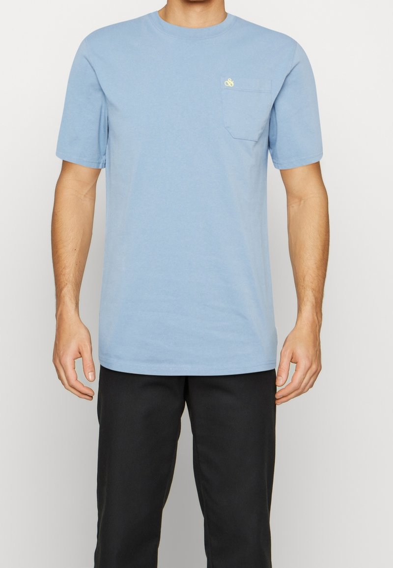 Vaaleansininen puuvillapaita, jossa on lyhyet hihat, pyöreä kaula-aukko ja rintatasku. Taskussa on pieni keltainen logo. Slim fit -malli.