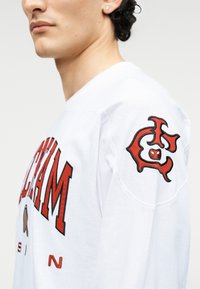 Λευκό πουκάμισο μακρυμάνικο με έντονα κόκκινα γράμματα στο μπροστινό μέρος και γραφική κηλίδα σε κόκκινο και μαύρο στον δεξί μανίκι.