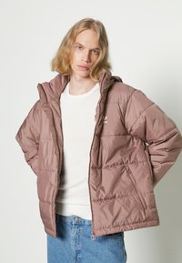 Giacca imbottita marrone con cappuccio, dotata di tasche con zip, design cucito e logo sul petto. Indossata sopra una camicia bianca e jeans blu.