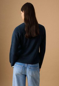 Femeie cu păr lung și întunecat purtând un pulover albastru închis și blugi albaștri deschis, stând cu fața către un fundal bej.