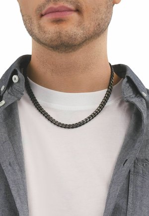 Nahaufnahme des unteren Gesichts eines Mannes, der eine schwarze Kettenhalskette, ein weißes T-Shirt und ein aufgeknotetes graues Hemd vor weißem Hintergrund trägt.