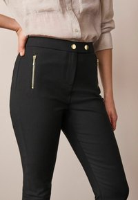 Zwarte, aansluitende broek met een gladde textuur, ritszakken en gouden knoopaccenten op de tailleband. Heeft een slanke silhouet.