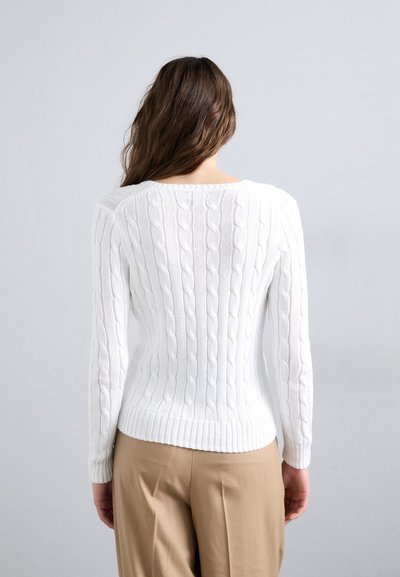 Pull en tricot blanc à mailles torsadées avec des manches longues et un ourlet côtelé, présentant des motifs texturés et un décolleté large. Associé à un pantalon beige.