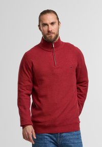 Roter, gestrickter Pullover mit Viertel-Zip und hohem Kragen sowie gerippten Bündchen. Besitzt eine subtile Textur und ein kleines Logo in der Nähe der Brust.
