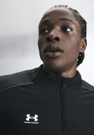 Close-up af en person med flettet hår iført en sort Under Armour lynlåstrøje til sport, der kigger til siden med et fokuseret udtryk.