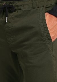Olijfgroene shorts met een gestructureerde stof, elastische tailleband en een zwart trekkoord. Voorzien van zijzakken met contrasterende binnenvoering.