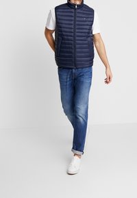 Mörkblå pufferväst med hög krage, framdragkedja och två sidofickor, bärs över en vit t-shirt och matchas med uppvikta jeanstights.