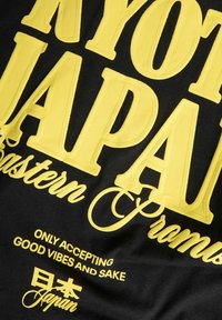Sort t-shirt med stort gult grafisk tekst: "KYOTO JAPAN," "Eastern Promise," og "ONLY ACCEPTING GOOD VIBES AND SAKE," med japanske tegn.