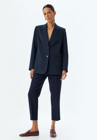Blazer navy à rayures fines avec deux boutons, revers crantés et poches avant associé à un pantalon tailleur navy et des mocassins bordeaux.