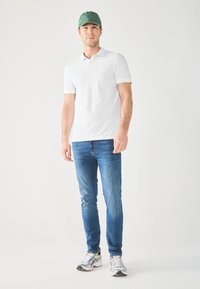Biely pólový tričko, modré džínsy a strieborné tenisky. Model má na hlave zelenú čiapku. Jednoduchý dizajn, hladká textúra a priliehavý strih.