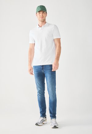 Weißes Poloshirt, blaue Jeans und silberne sneakers. Model trägt eine grüne Kappe. Einfaches Design, glatte Textur und taillierte Form.