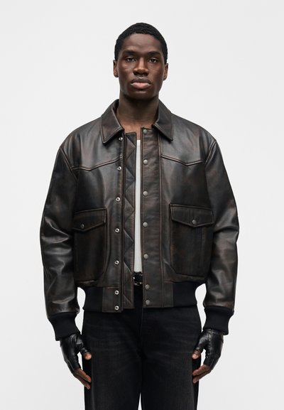 Καφέ δερμάτινο μπουφάν bomber με μπροστινές τσέπες, κουμπιά με κλιπ, ριμπ μανσέτες και λεία επιφάνεια. Φορεμένο πάνω από λευκό πουκάμισο και μαύρα τζιν.