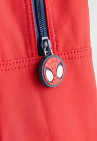 Tela roja con acentos cosidos, que presenta un tirador de cremallera azul marino y un charm redondo temático de Spiderman con ojos blancos y un patrón de telaraña roja.